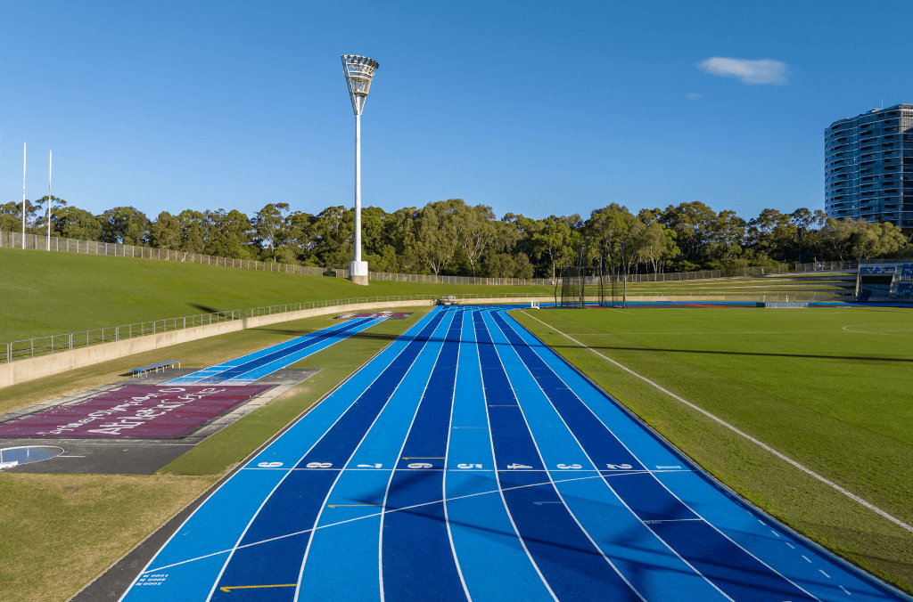 Blue running track with marked lanes stretching through a green athletics field, bordered by grassy embankments, trees, and a tall floodlight tower.