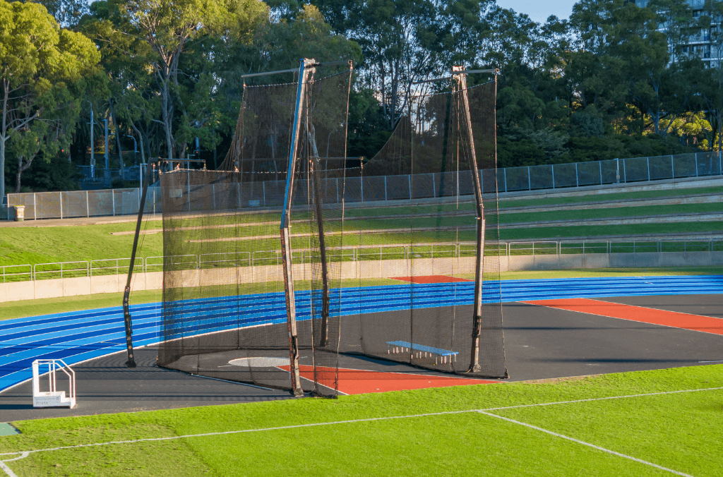 Caged throwing area for discus or hammer with tall netting stands beside a blue running track and green field, surrounded by trees and grassy terraces.