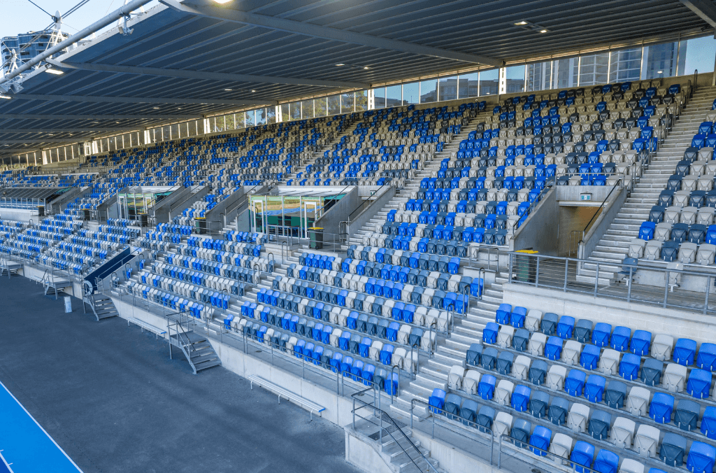 Covered grandstand with rows of blue and grey stadium seats overlooking an athletics track, featuring stairways, railings, and enclosed viewing areas beneath a steel roof.