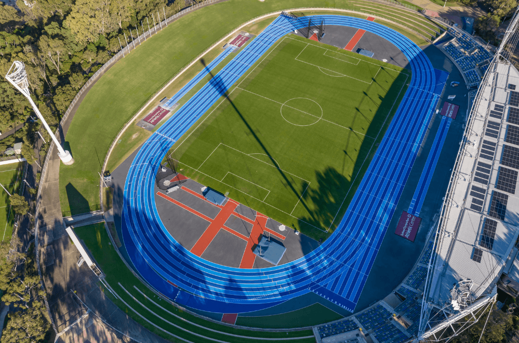 Aerial view of a blue athletics track encircling a green sports field with marked lanes and throwing areas, bordered by trees, seating, and floodlight towers.