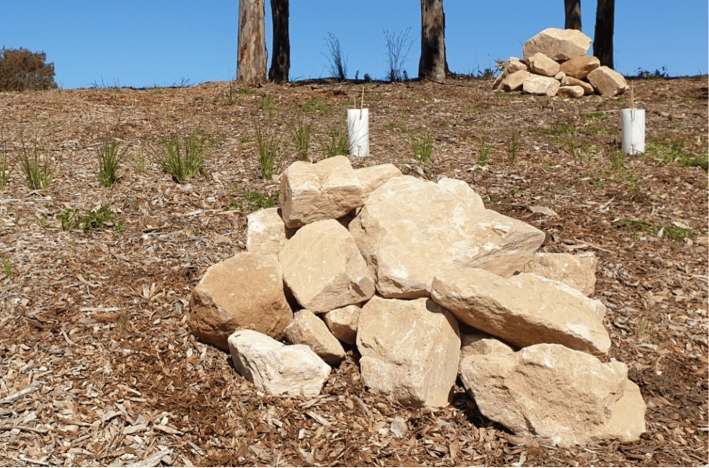 Pile of pale sandstone rocks on a mulched hillside with newly planted native grasses and trees under a clear blue sky.